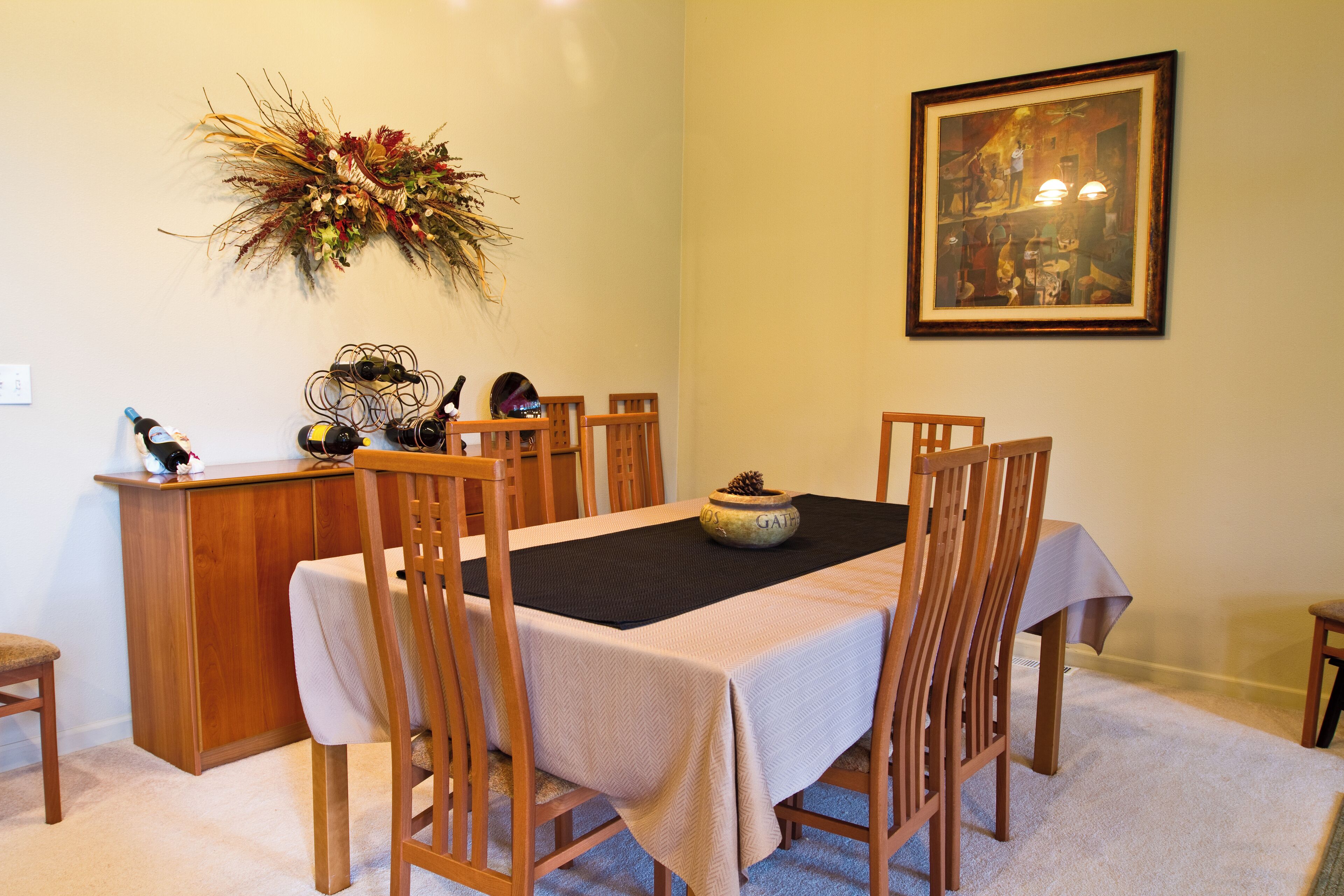 Zona de dinar amb una taula preparada per a sis, envoltada d'amb cadira de fusta. Un armari de fusta amb una ampolla de vi i gots està a l'esquerra. Un ram decoratiu penja a la paret sobre la taula.
