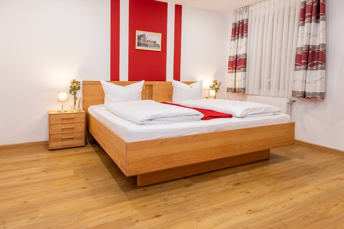 Un lit double bien fait, avec du linge de lit blanc, une tête de lit en bois et un plaid rouge. Il y a une table de chevet avec une lampe et un petit vase de fleurs. La pièce a un plancher en bois et un mur rayé de rouge et de blanc.