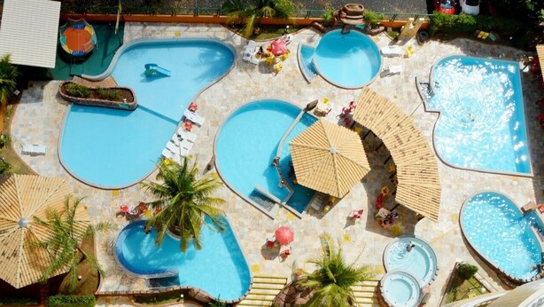 A large outdoor swimming pool area with multiple pools, sun loungers, umbrellas, and palm trees.