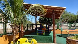 A playground with slides and climbing structures, surrounded by a fence and palm trees.