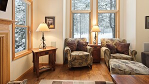 A cozy living room with a fireplace, two armchairs, a wooden coffee table, and a large window with a view of a snowy forest. - 9 Hoodoo Home with XBOX 360 and Private Hot Tub by RedAwning (Bend)