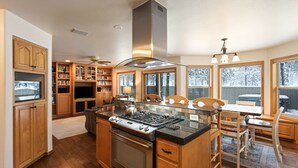 A modern kitchen with wooden cabinets, a central island with a stove, and large windows offering views of the outdoors. - 9 Hoodoo Home with XBOX 360 and Private Hot Tub by RedAwning (Bend)