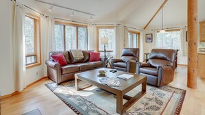 A spacious living room with a brown leather sofa, two leather armchairs, a wooden coffee table, and a patterned rug. Large windows allow natural light to fill the room. - 7 Cedar Private Cul-de-sac include Hot Tub with Forest View by RedAwning (Bend)