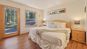 A bedroom with a large bed, wooden floors, a view of a forest through a sliding door, and a small window. - 1 Colonial Private Family Vacation Home features Bikes to Explore Sunriver by RedAwning (Bend)