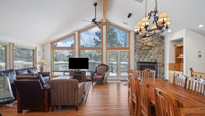 A spacious living room with a large wooden dining table, comfortable seating, a fireplace, and large windows offering a view of the outdoors. - 2 Sarazen Home with Hot Tub and BBQ on the Deck, Close to Woodlands Golf Course by RedAwning (Bend)