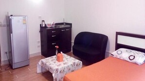 Una habitación pequeña y sencilla con cama, una silla, una mesa, refrigerador y cómoda.