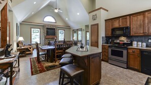 A spacious kitchen with a stone fireplace, wooden cabinets, and a large island with bar stools. - Elite Escape-2 POOLS, Splash Pad, PoolTable, Hot Tub, Patio w/Fire Pit+Fireplace (Hollister)
