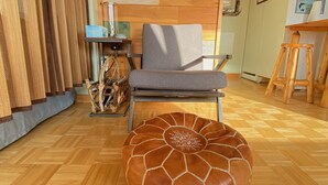 A room with a wooden herringbone floor, a grey armchair, a round ottoman with a geometric design, a wooden wall panel, and a clock on the wall. - Relaxing Elevated Lake House Retreat (Mayville)