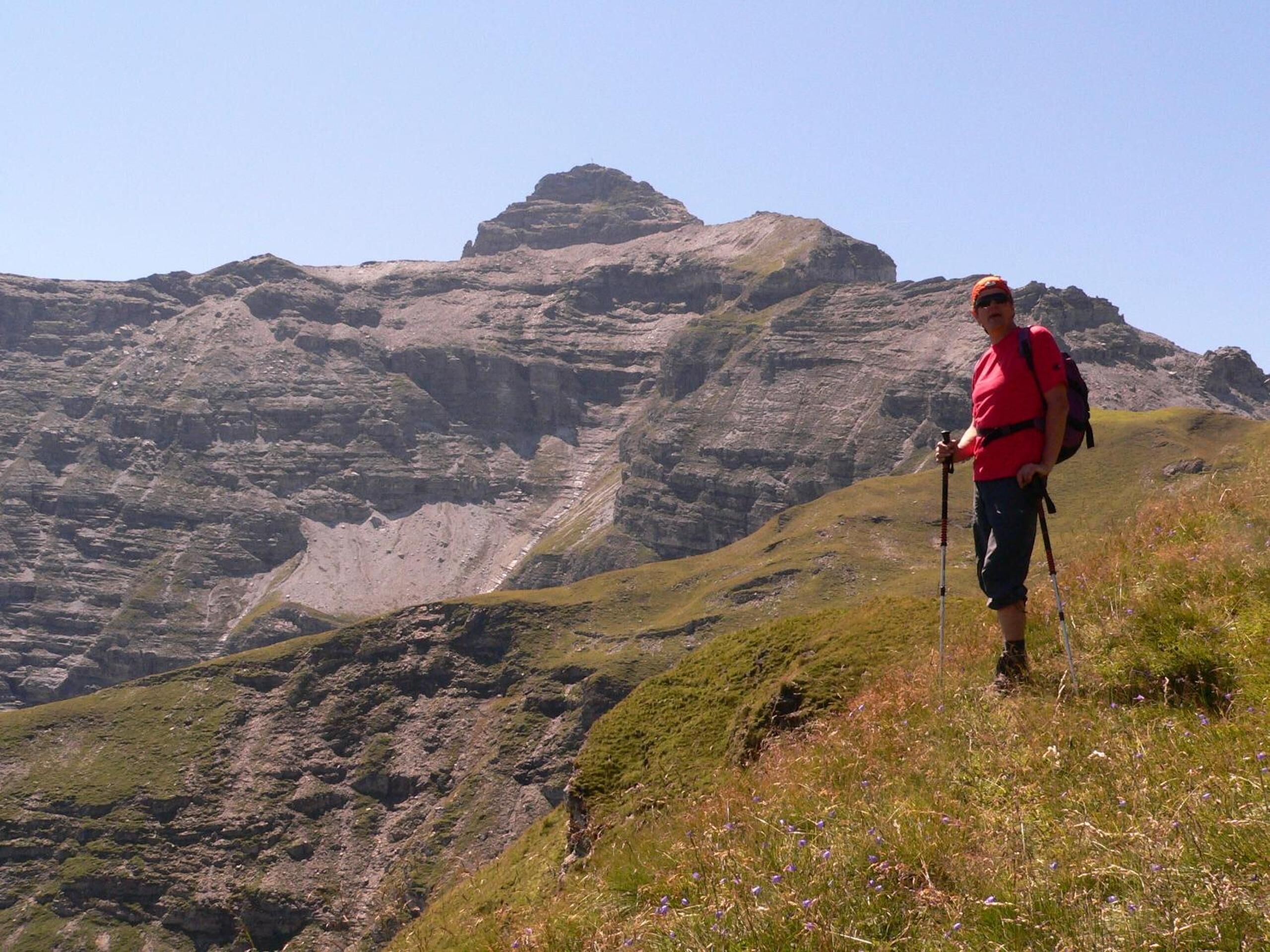Ein Wanderer auf einem grasbewachsenen Berghang mit felsigem Untergrund und einem markanten Gipfel im Hintergrund.