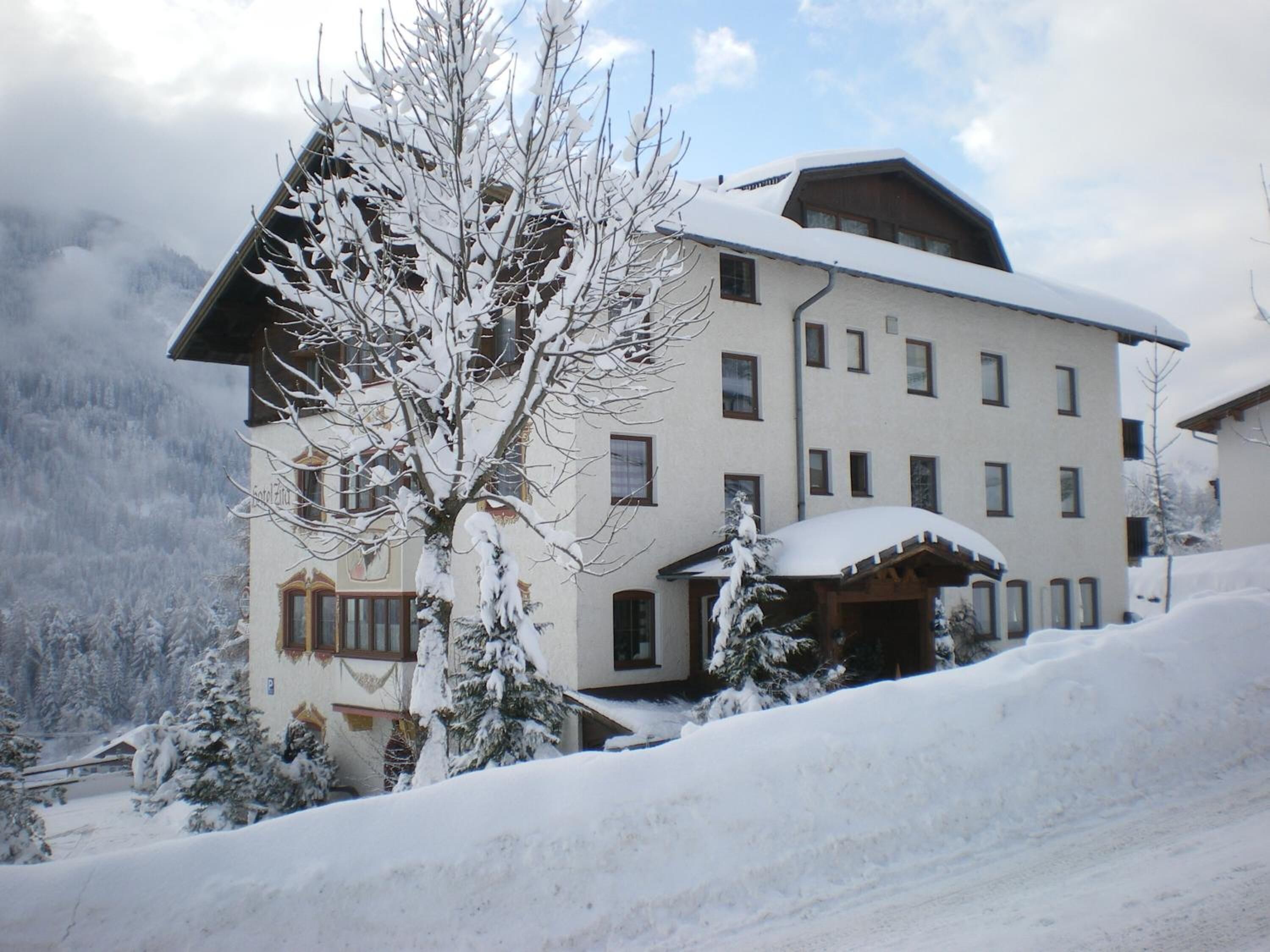 A multi-story hotel with a snow-covered exterior, a covered entrance, and a mountainous backdrop.