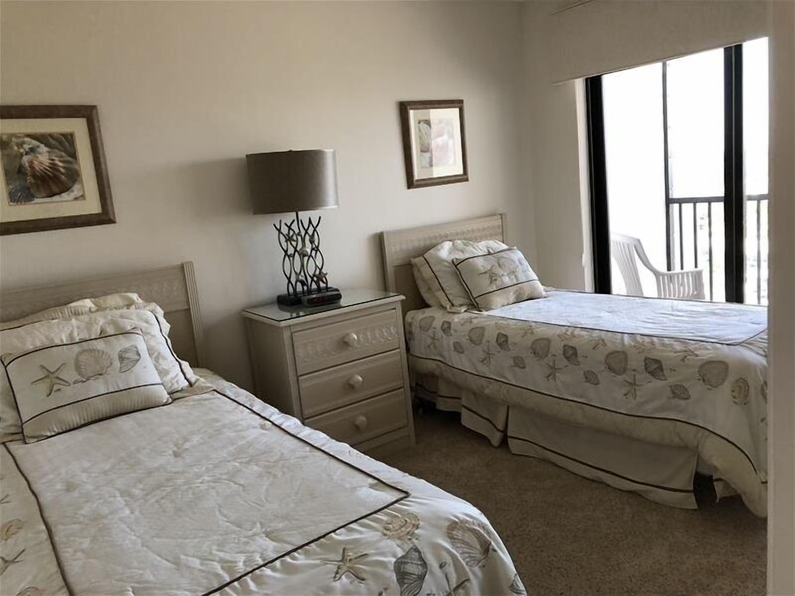 A bedroom with two beds, a nightstand, a lamp, and a window with a view.