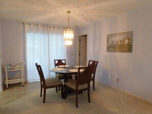 A dining room with a round table, four chairs, a chandelier, a painting on the wall, and a small white shelf with items on it.
