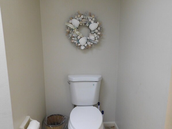 A toilet with a white seat and tank, a wall-mounted decorative wreath, and a trash can.