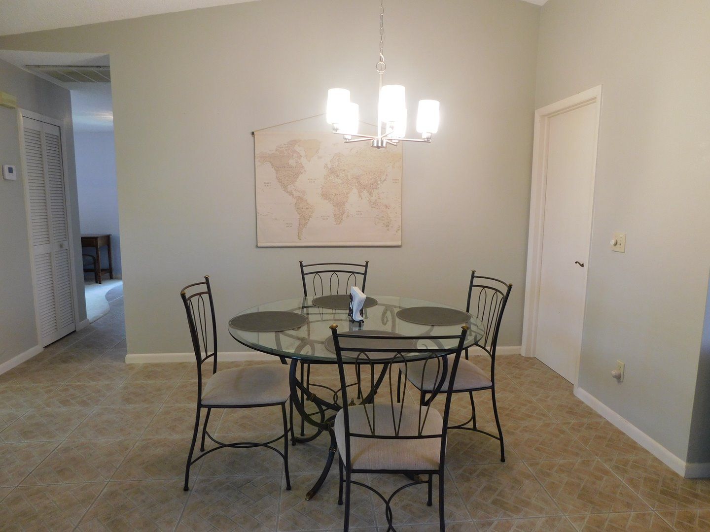 A dining area with a glass table, four chairs, a world map on the wall, and a chandelier.