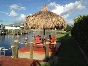 Un espacio de estar al aire libre con techo de paja, terraza de madera y sillas, con vista a una masa de agua con un muelle y edificios al fondo.