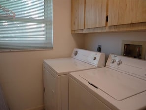 A laundry room with two white washers and dryers, wooden cabinets above, and a window with blinds.