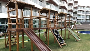 A wooden playground structure with slides and climbing elements, surrounded by a well-maintained lawn and a pool in the background. - Beautiful luxury apartment, 3 suites, 55" TV, swimming pools, air conditioning, complete, 400m from the beach, cond. (Aquiraz)