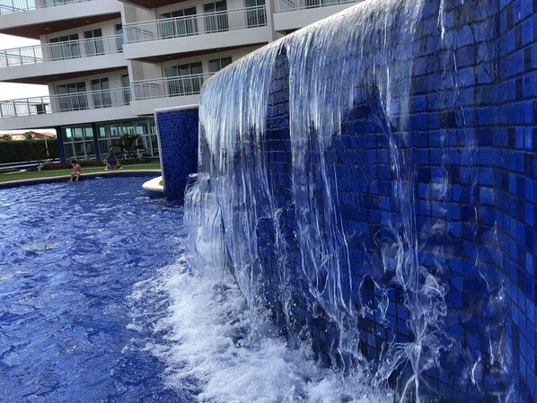 A large indoor water feature with a cascading waterfall, surrounded by a pool. - Beautiful luxury apartment, 3 suites, 55" TV, swimming pools, air conditioning, complete, 400m from the beach, cond. (Aquiraz)
