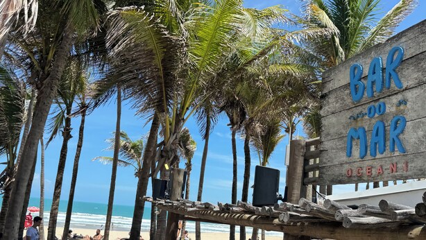 A beach bar with a wooden sign, palm trees, and beachgoers. - Beautiful luxury apartment, 3 suites, 55" TV, swimming pools, air conditioning, complete, 400m from the beach, cond. (Aquiraz)