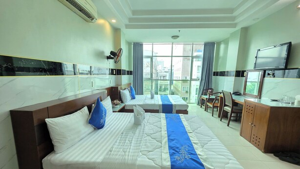A hotel room with two beds, a desk, a TV, and a balcony with a city view.
