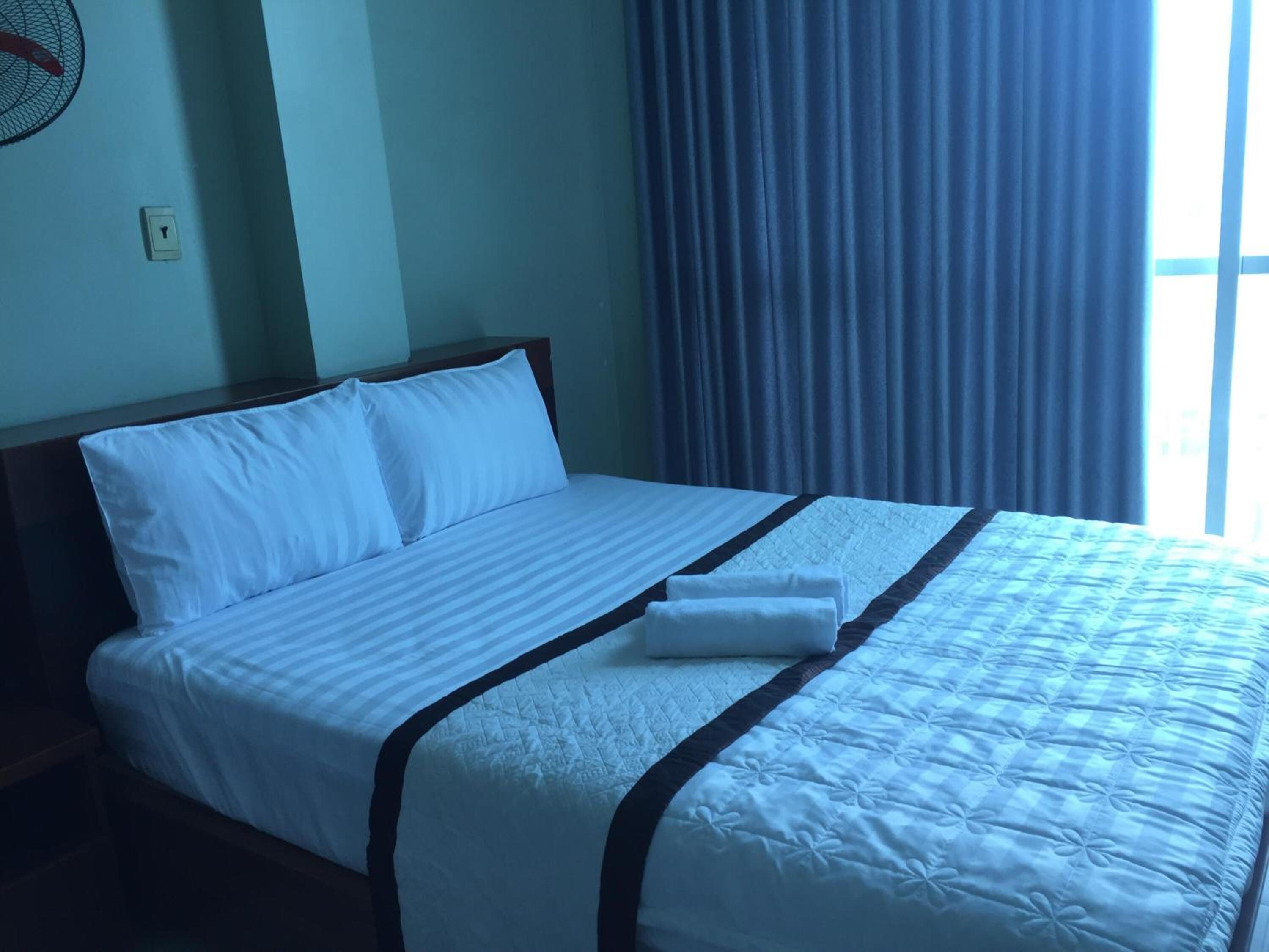 A neatly made bed with white bedding and a dark headboard, a folded towel on the bed, and a window with blue curtains.