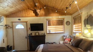 Une chambre confortable avec un plafond en bois, un lit recouvert d’une couverture marron, un bureau avec une lampe et un téléviseur fixé au mur.