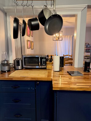 A kitchen with a microwave, toaster, blender, and hanging pots. There is a wooden cutting board, a paper towel holder, and a tablet on the counter.