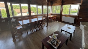 A rustic wooden dining area with a long table and benches, large windows with views of greenery, and a radiator on the wall.