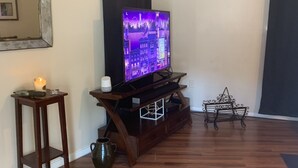 Ett rum med trägolv, en tv på ett tv-bord, en stol, ett litet bord med en lampa och en spegel som hänger på väggen.