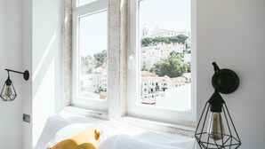 Une chambre à coucher avec un lit, une fenêtre donnant sur des immeubles et un luminaire fixé au mur.