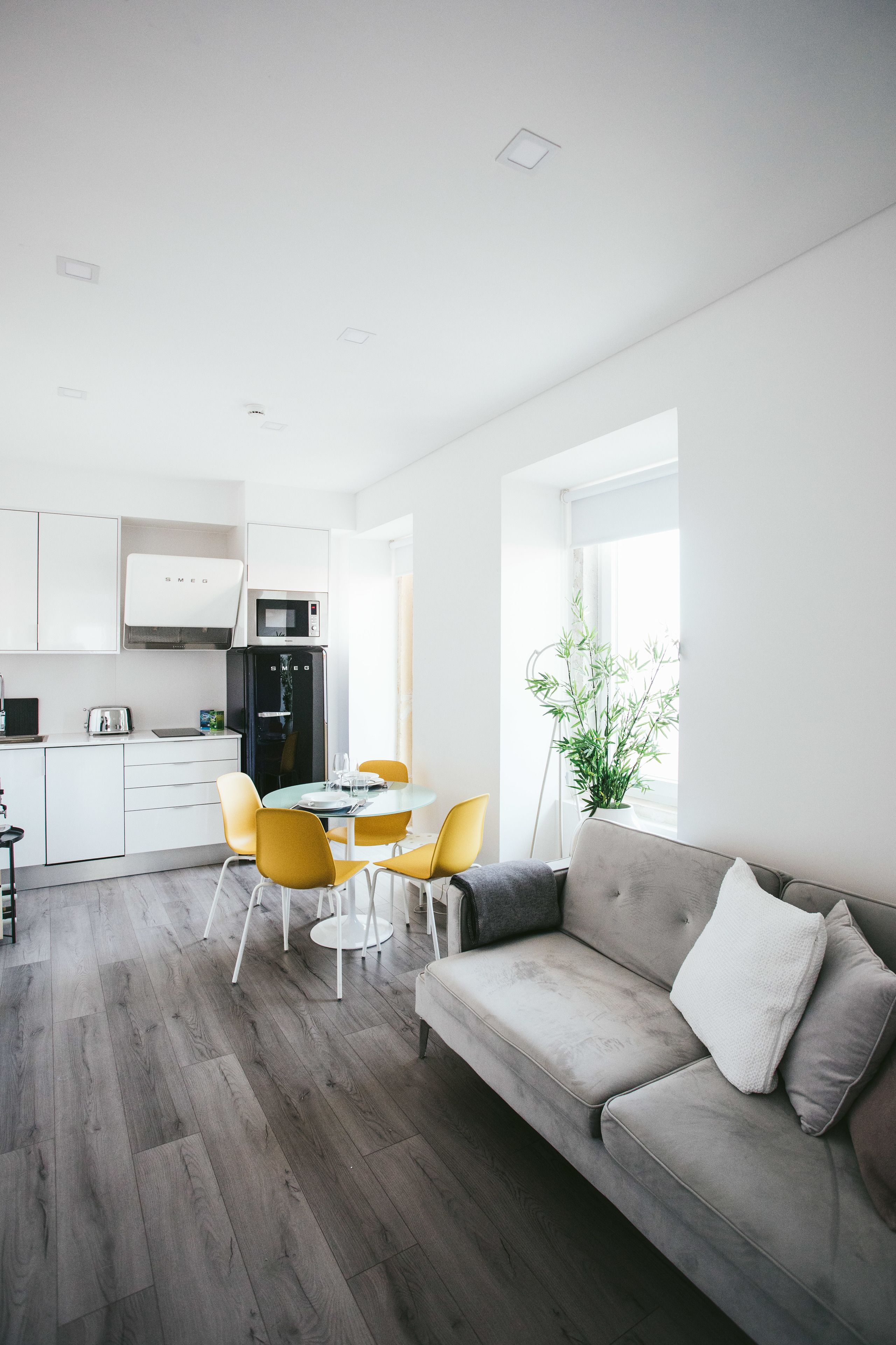 A modern kitchen with white cabinets, a microwave, and a refrigerator. There is a dining table with yellow chairs and a gray sofa with cushions.