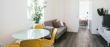 A modern living room with a glass dining table, yellow chairs, a grey sofa with cushions, and a large potted plant.