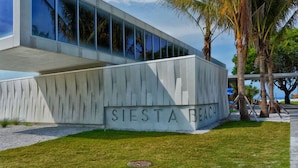 Un bâtiment moderne dont la façade porte l’inscription « SIESTA BEACH », entouré de palmiers et d’une pelouse.