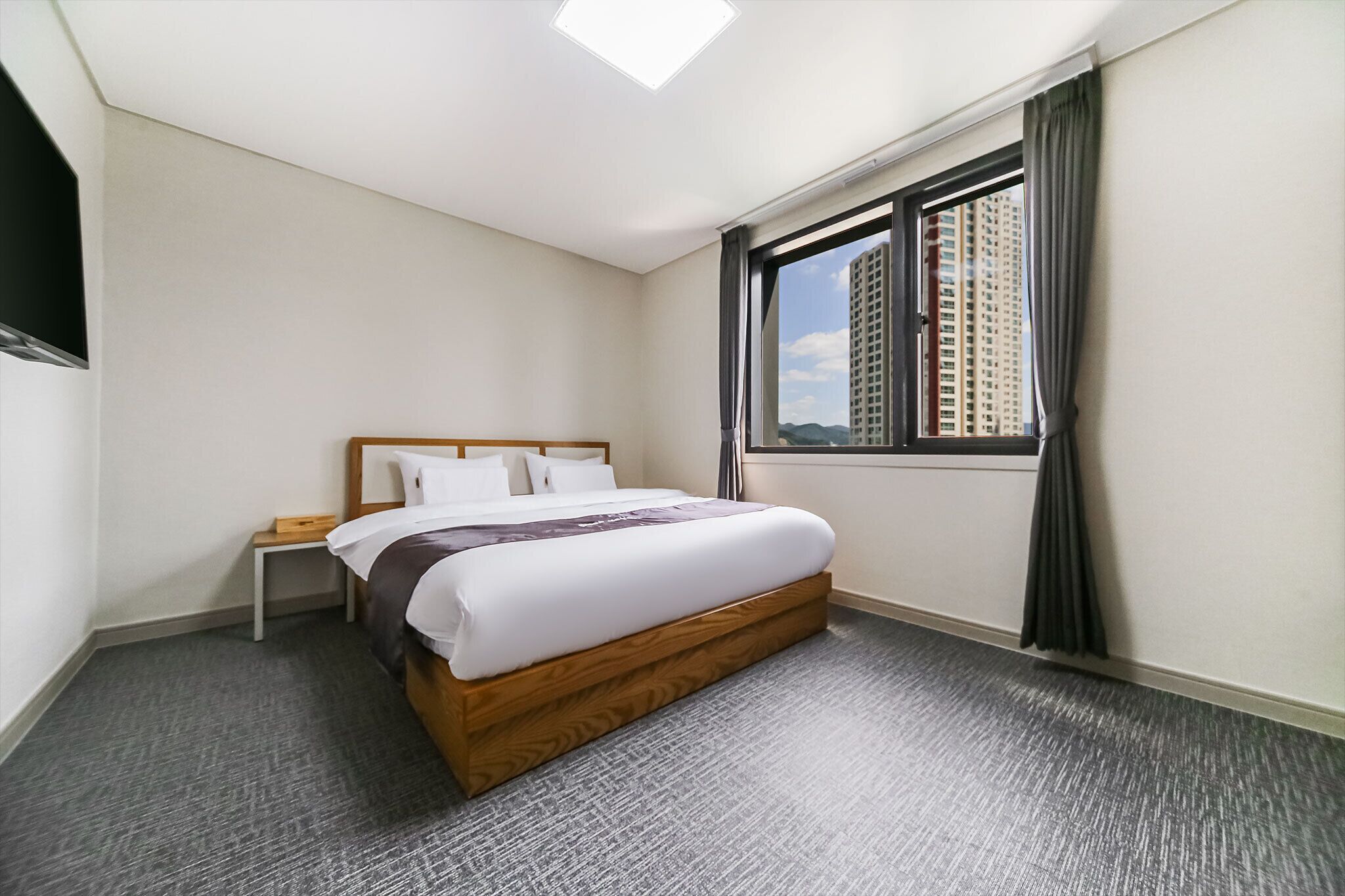 A hotel room with a large bed, a nightstand, a window with curtains, and a view of tall buildings.