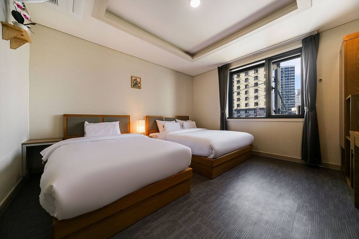 A hotel room with two beds, a window with a city view, a small painting on the wall, and a wooden wardrobe.
