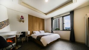 A hotel room with a bed, a small table with a chair, a fireplace, and a window with a view of buildings. - Masan Amor Hotel (Changwon)