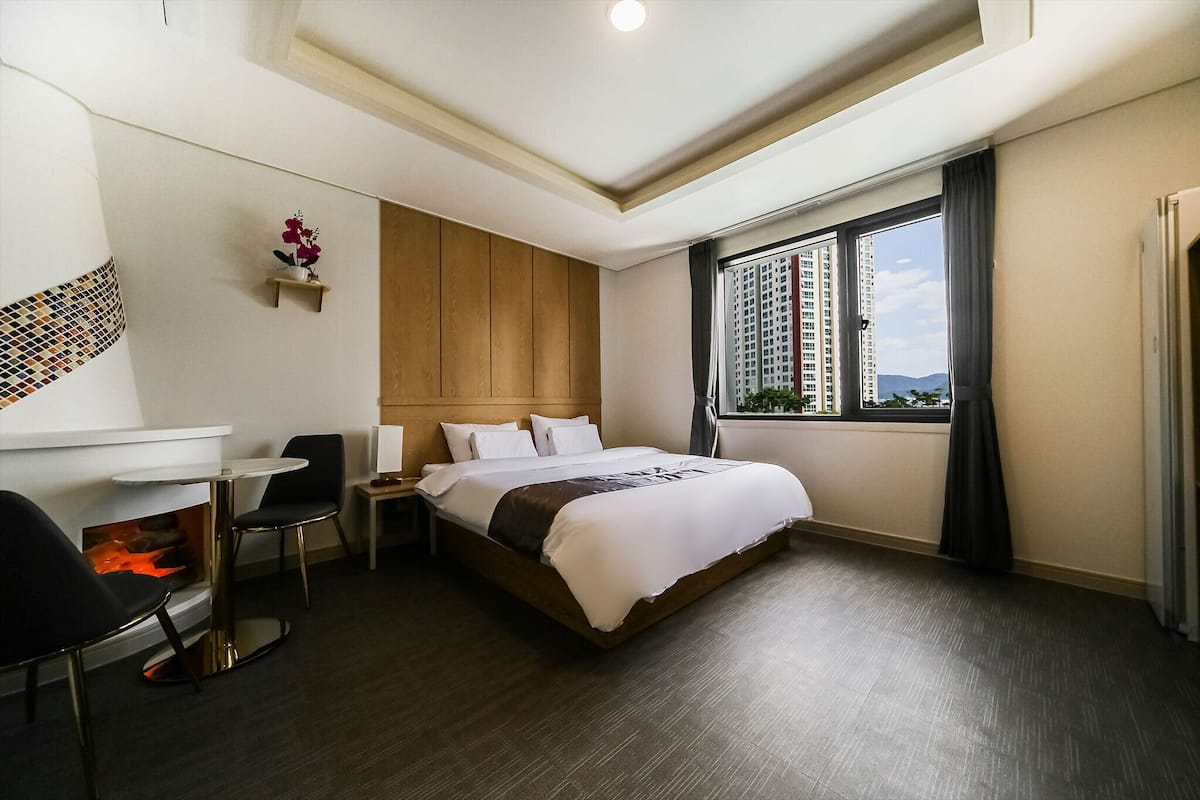 A hotel room with a bed, a small table with a chair, a fireplace, and a window with a view of buildings.