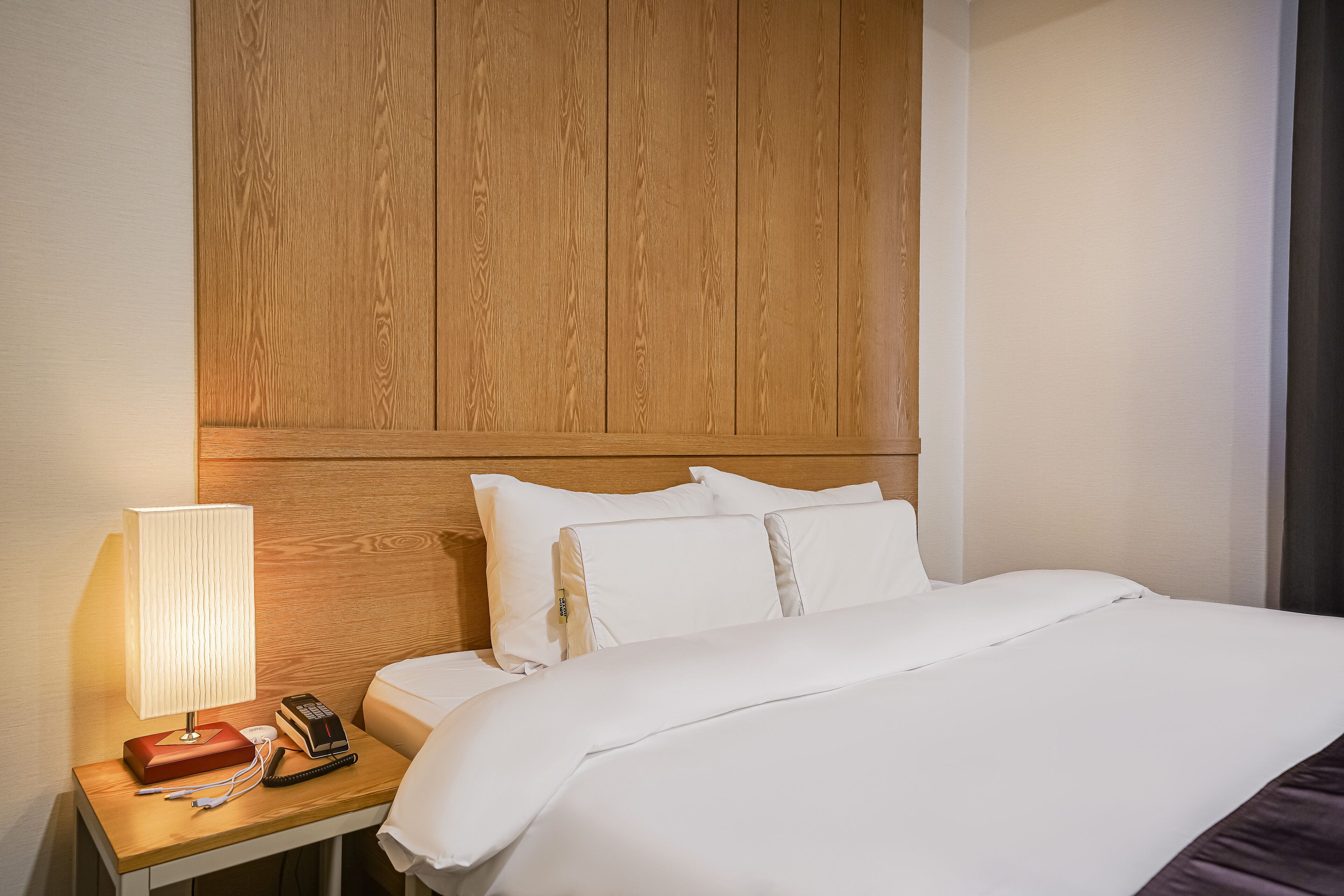 A hotel room with a wooden headboard, a bedside table with a lamp, a telephone, and a neatly made bed with white bedding.