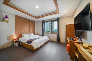 Ein modernes Hotelzimmer mit einem großen Bett, einem Schreibtisch mit Computer, einem Fernseher und einem Fenster mit Blick auf die Stadt.