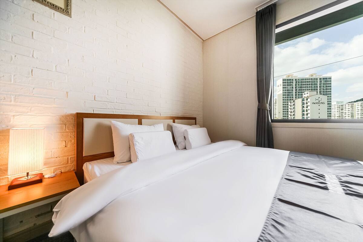 A hotel room with a white brick wall, a wooden headboard, a bed with white bedding, a nightstand with a lamp, and a window with a view of buildings.