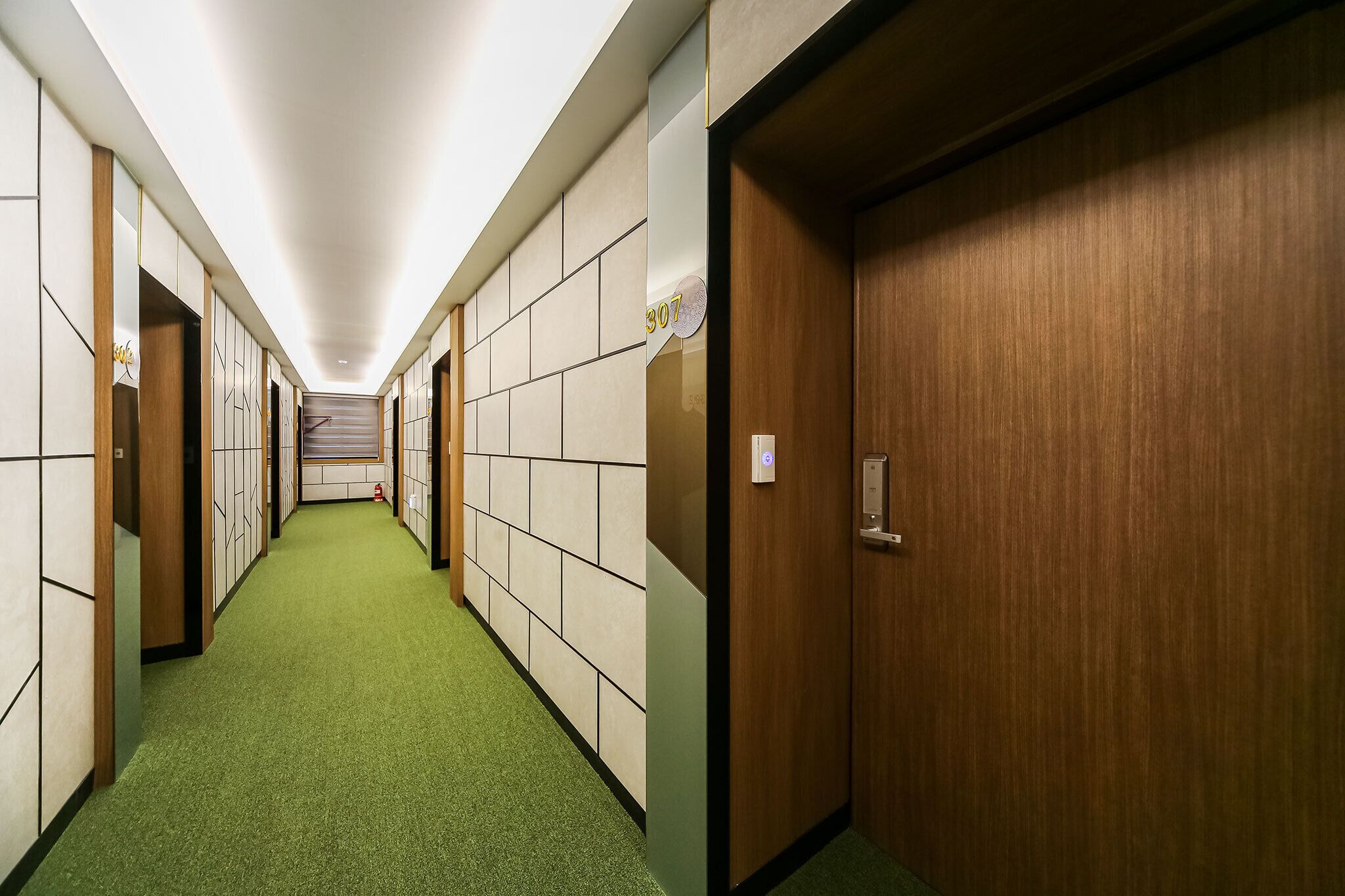Un corridoio d'albergo con tappeto verde, porte in legno e parete rivestita di piastrelle.