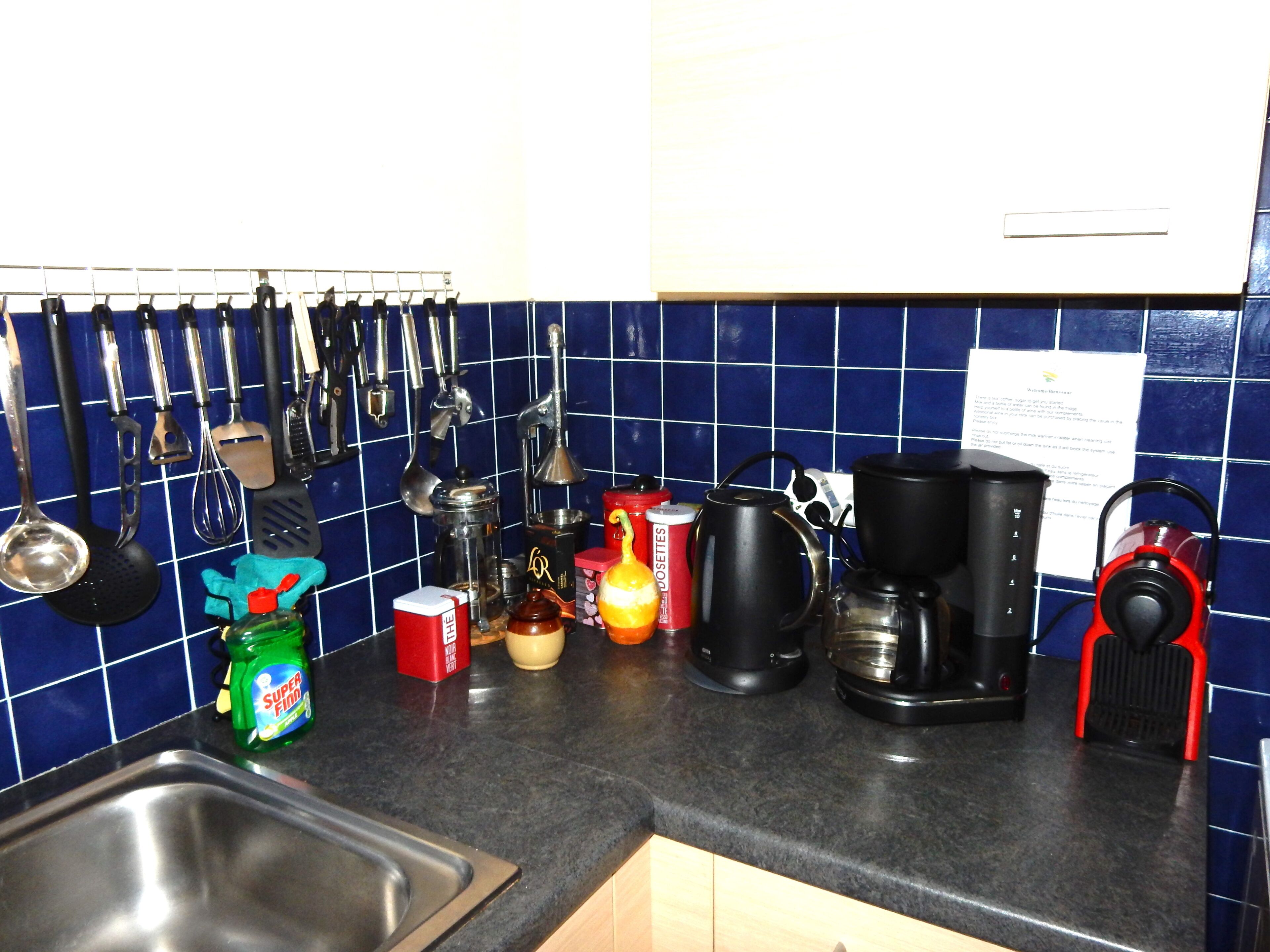Et køkken med en vask, diverse køkkenredskaber, en elkedel, en kaffemaskine og en rød elkedel på bordet.