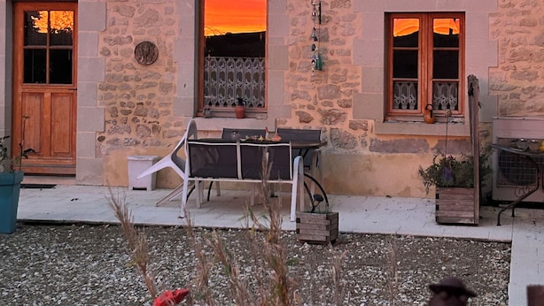 A stone building with a large window reflecting a sunset, a door, and a small table with chairs outside.