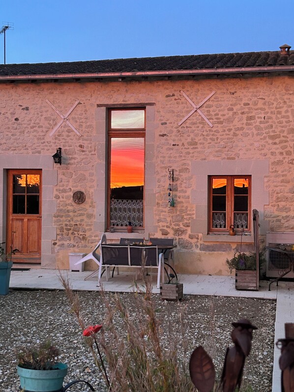 A stone building with a large window reflecting a sunset, a door, and a small table with chairs outside.