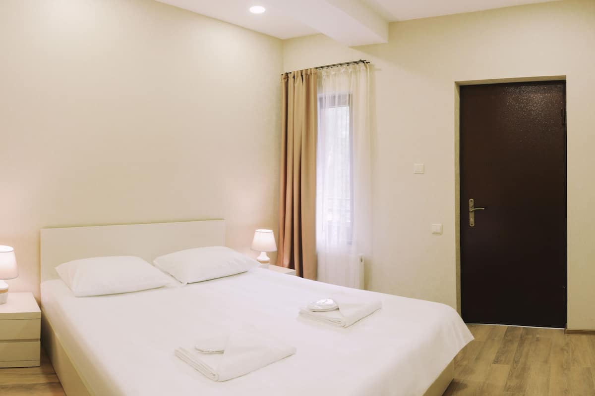 Cama bem arrumada com roupa de cama branca, duas luminárias de cabeceira, uma porta escura e uma janela com cortinas bege.
