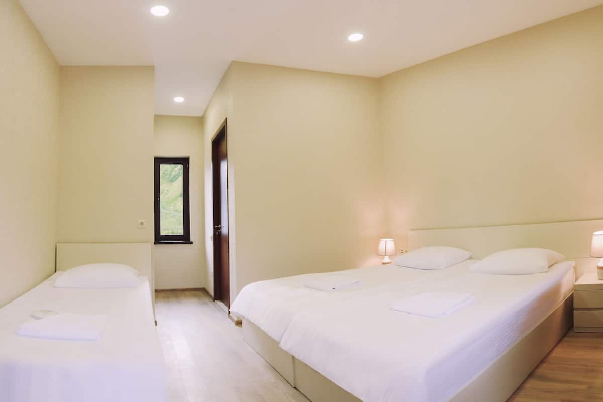 Quarto de hotel com duas camas de solteiro, uma mesa de cabeceira com luminária e uma porta que dá para outro cômodo.