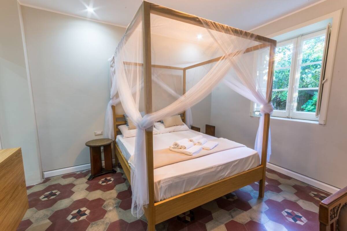 A four-poster bed with a canopy, a bedside table, and a window with a view of greenery.