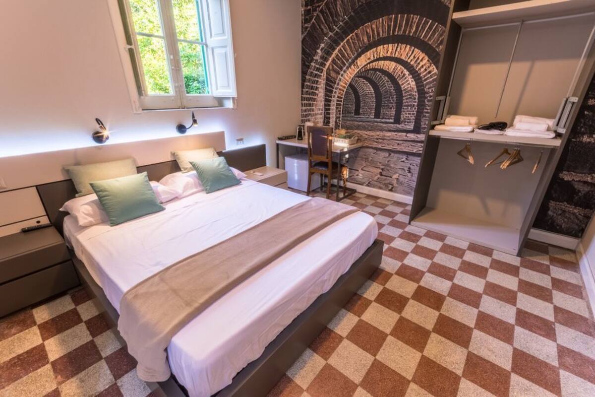 A bedroom with a checkered floor, a bed with pillows, a desk with a chair, a stone wall, and a wardrobe.