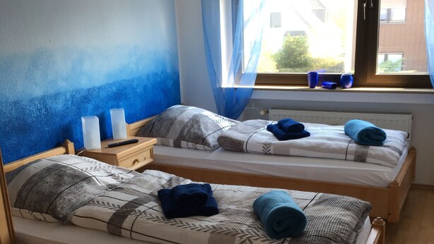 Ein Zimmer mit zwei Holzbetten, jeweils mit Kissen und Decke. An den Betten stehen Nachttische mit Lampen, und ein Fenster mit Vorhängen.