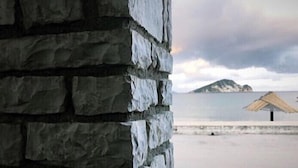 A stone wall with a view of a beach and a thatched umbrella. - Villa Miranda (Zakynthos)